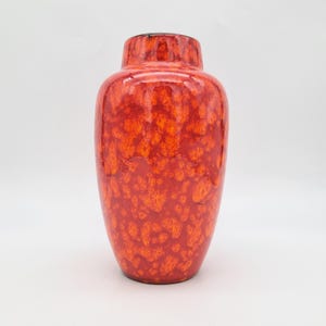 Puede incluir: Un jarrón de cerámica con acabado brillante que presenta un patrón moteado en rojo y naranja vibrante. El recipiente tiene un cuerpo redondeado que se estrecha hacia un cuello angosto y una abertura circular en la parte superior.