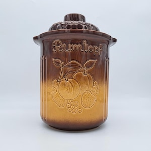 Könnte beinhalten: Ein brauner Rumtopf-Topf aus Keramik mit Deckel. Der Topf zeigt geprägte Obstmotive, darunter Pfirsiche, Erdbeeren und Trauben. Das Wort "Rumtopf" steht oben. Der Topf hat ein geripptes Design und eine zweifarbige braun-beige Farbgebung.