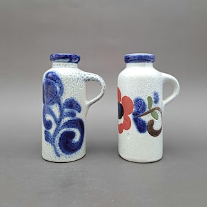 Peut inclure: Deux vases en céramique avec poignées. L'un présente un motif floral bleu, l'autre une fleur rouge et bleue avec des accents verts et bruns. Les deux ont un fond blanc moucheté texturé et des bords bleus.
