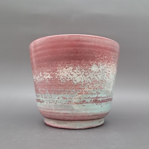 May include: A handmade ceramic planter with a speckled finish. The planter features a gradient of colors, transitioning from a deep red at the top to a light blue-green at the base. The planter has a cylindrical shape with a slightly flared rim.