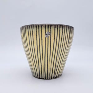 Pode incluir: Um vaso de cerâmica com uma cor de base amarelo-pálido e riscas verticais castanho-escuras que se estreitam em direção à base. O recipiente tem um topo largo e aberto e um pequeno autocolante prateado circular com um logótipo preto.