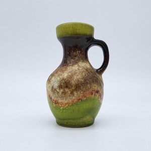 Puede incluir: Un pequeño jarro de cerámica con asa. La parte superior es verde oliva, que pasa a marrón oscuro, con un cuerpo moteado beige y marrón. La mitad inferior es de un verde vibrante. El jarro tiene forma redondeada y un asa curva.