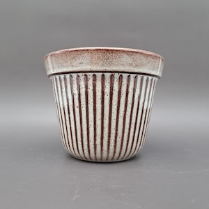 Puede incluir: Una maceta de cerámica con base crema y rayas verticales marrones. El borde es de color marrón oscuro. La maceta tiene forma cilíndrica y es adecuada para uso en interiores o exteriores. Los colores neutros la hacen versátil.