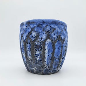 Op de afbeelding: Een handgemaakte keramische beker met een uniek, getextureerd oppervlak. De beker heeft een opvallende blauwe glazuur met donkere, bijna zwarte accenten die een visueel interessant patroon creëren. Het ontwerp is organisch en de beker heeft een licht onregelmatige vorm.