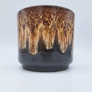 Puede incluir: Una maceta cilíndrica de cerámica con una base marrón oscuro y una sección superior con un efecto de esmalte goteado en tonos marrón, beige y blanco. La maceta tiene un borde ligeramente ensanchado y un acabado liso y brillante.