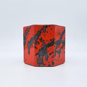 Puede incluir: Un recipiente cerámico hexagonal rojo con diseño de pintura negra salpicada. El recipiente tiene un acabado brillante y está sobre un fondo blanco. El diseño le da al recipiente un aspecto moderno y artístico.