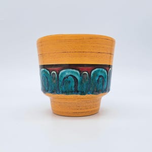 Peut inclure: Un cache-pot en céramique avec une section supérieure et inférieure orange vif dotée d'une finition texturée à coups de pinceau horizontaux. La bande centrale présente un motif décoratif d'arches bleu sarcelle et bleu foncé sur fond rouge.