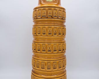 May include: Tall, cylindrical ceramic vase in shades of brown and tan. The vase features a series of stacked, rounded bands, each adorned with a repeating geometric pattern. The vase has a wide base and a narrower top.