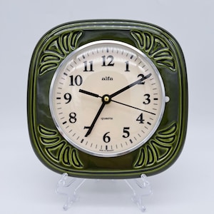 May include: A vintage green quartz clock with a square, decorative frame. The clock face is cream-colored with black numerals and hands. The frame has an embossed leaf design. The clock is on a clear plastic stand.