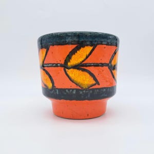 Könnte beinhalten: Ein Keramik-Pflanztopf mit einem lebendigen Design. Der Topf hat einen leuchtend orangefarbenen Boden und Körper mit einem schwarzen Band oben und unten. Der orangefarbene Abschnitt ist mit gelben und orangefarbenen Blättern verziert. Der Rand ist dunkelblau.