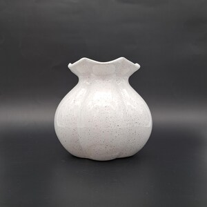 Könnte beinhalten: Eine weiß gesprenkelte Keramikvase mit gerüschtem Rand. Die Vase hat einen abgerundeten Körper und ein gesprenkeltes Muster, das ein strukturiertes Aussehen erzeugt. Die Vase steht vor einem dunklen Hintergrund.