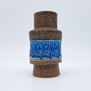 以下が含まれることがあります： 茶色のセラミック花瓶。表面はざらざらしており、円筒形です。中央には青色の模様のあるセラミックの帯があります。花瓶は底が広く、上部が細くなっています。