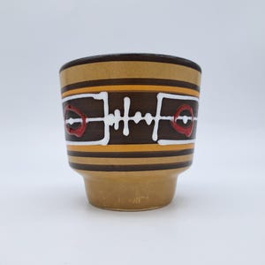 Op de afbeelding: Een keramische mok met horizontale banden in bruin en mosterdgeel. Het midden is versierd met een wit lineair geometrisch patroon en twee rode cirkelvormige accenten. De mok heeft een brede, cilindrische bovenkant en een taps toelopende voet.