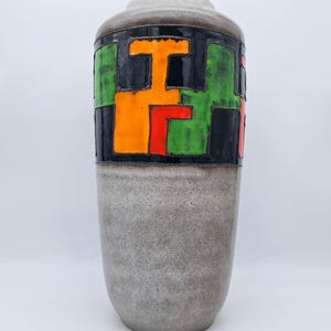 May include: A tall, gray ceramic vase with a geometric design. The upper portion features a black band with orange, green, and red shapes. The vase has a textured surface and a rounded top.
