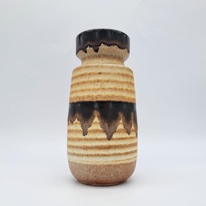 Puede incluir: Un jarrón de cerámica de forma cilíndrica, con bandas horizontales en tonos beige y marrón. La parte superior y una sección central son de color marrón oscuro, con un diseño texturizado y acanalado. La base es marrón claro.