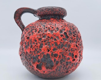 May include: A red and black ceramic vase with a textured, volcanic-like surface. The vase has a rounded body, a small opening, and a curved handle. The color scheme is primarily red with black accents, creating a striking contrast.