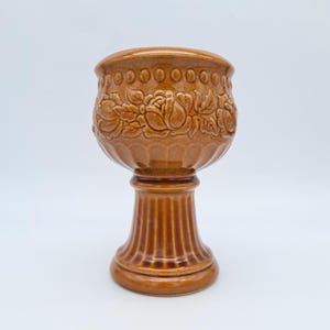 May include: A glossy brown ceramic goblet featuring a raised relief pattern of blooming roses around the bowl. The upper rim is decorated with a series of small circular indentations, and the base is supported by a thick, vertically fluted pedestal.