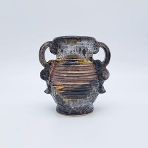 Puede incluir: Un pequeño jarrón de cerámica hecho a mano con dos asas. El jarrón presenta una superficie texturizada con una paleta de colores marrón, negro y dorado. El diseño incluye bandas horizontales y elementos decorativos cerca de las asas, creando una estética rústica.