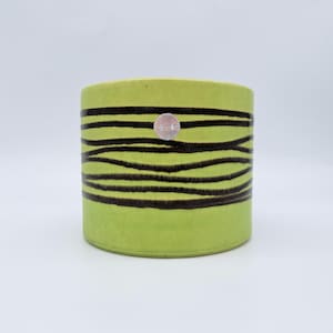 May include: A cylindrical ceramic planter in a bright lime green colour featuring horizontal, wavy dark brown stripes wrapping around the exterior. A small, circular silver sticker is affixed to the upper centre of the outer surface.