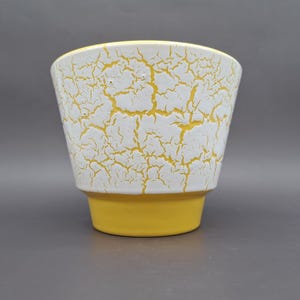 Op de afbeelding: Een keramische plantenpot met een wit bovengedeelte met een gele craquelé afwerking en een effen gele basis. De pot heeft een taps toelopende vorm met een brede rand en een gladde, platte bodem, wat het contrast tussen het textuurrijke craquelé-oppervlak en het effen gele onderste gedeelte benadrukt.