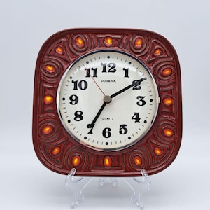 May include: A square, vintage-style clock with a burgundy frame and a cream-coloured face. The clock face features black numerals and hands, with the word "DUGENA" above the centre. The frame has an ornate design with orange accents.