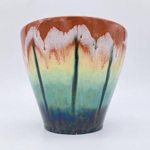 Op de afbeelding: Een keramische plantenpot met een uitlopende rand en een reactief glazuur dat verloopt van diep donkerblauw aan de basis naar lichtgroen, geel en uiteindelijk roodbruin aan de bovenkant, met donkere verticale druippatronen op het oppervlak.