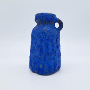 Pode incluir: Um pequeno jarro de cerâmica cilíndrico com um esmalte azul vibrante e uma superfície porosa fortemente texturizada. O recipiente apresenta um gargalo curto e estreito, uma pequena alça circular na lateral e uma base plana com acabamento rugoso.