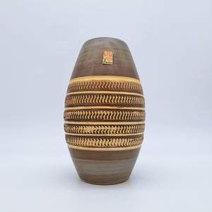 Puede incluir: Un jarrón de cerámica marrón con un diseño texturizado y estampado. El jarrón tiene una forma redondeada con un cuello estrecho y una base ancha. El diseño presenta bandas horizontales de patrones repetidos en tonos marrones y dorados. Una pequeña etiqueta está adherida al jarrón.