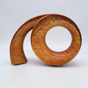 Könnte beinhalten: Eine Keramikskulptur mit einem Spiraldesign. Die Skulptur hat eine warme braune Farbe mit einer strukturierten Oberfläche und einer kreisförmigen Öffnung. Das Stück hat eine gebogene, längliche Form und ist wahrscheinlich ein Dekorationsartikel.