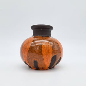 Könnte beinhalten: Kleine, runde Keramikvase mit dunkelbraunem Rand und Boden. Der Körper der Vase ist orange mit vertikalen schwarzen Streifen. Die Vase hat eine glänzende Oberfläche und steht auf einer weißen Oberfläche.