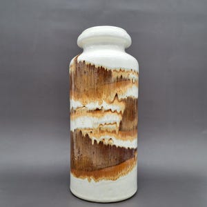 Puede incluir: Un jarrón cilíndrico de cerámica con tapa y base blancas, con un diseño texturizado de bandas horizontales marrones y beige. El jarrón tiene una parte superior redondeada y un acabado liso y brillante.