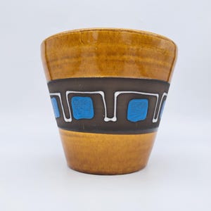 Könnte beinhalten: Ein Keramik-Pflanzgefäß mit glänzend brauner Oberfläche. Ein dunkelbraunes Band umschließt die Mitte mit einem weißen und blauen geometrischen Design. Der Pflanztopf hat eine leicht ausgestellte Form.