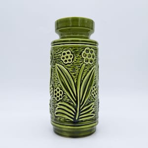 May include: A green ceramic vase with a cylindrical body and a flared top. The vase features a raised floral design with leaves and flower-like shapes. The color is a deep, earthy green, and the texture appears to be slightly rough.
