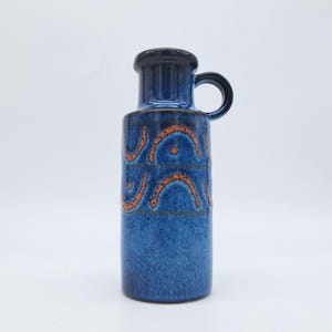 Könnte beinhalten: Eine blaue Keramikvase mit Henkel und orangefarbenem Wellenmuster. Die Vase hat einen dunkelblauen Rand und Henkel sowie einen zylindrischen Körper. Das orangefarbene Muster ist ein sich wiederholendes Design. Die Vase ist etwa 25 cm hoch.
