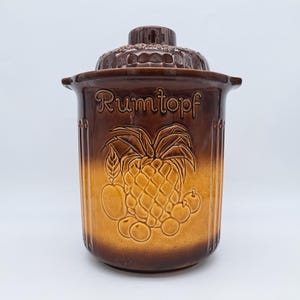 Könnte beinhalten: Brauner Keramik-Rumtopf mit Deckel. Der Topf zeigt ein Ananas- und Fruchtmuster mit dem Wort "Rumtopf" in Schrift. Der Topf hat ein zweifarbiges Farbschema, mit dunklerem Braun oben und hellerem Braun unten.