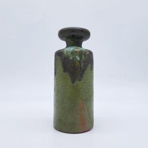 Può includere: Un vaso cilindrico in ceramica con una parte superiore arrotondata marrone scuro e un corpo verde. Il verde ha un aspetto screziato con accenti marroni più scuri vicino alla parte superiore. Il vaso ha un design semplice ma elegante.