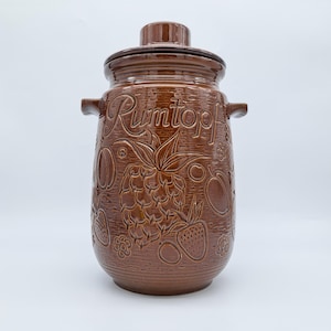 May include: A brown ceramic Rumtopf pot with a lid, handles, and embossed fruit designs. The pot features a pineapple, strawberries, and other fruits. The word "Rumtopf" is embossed on the front.