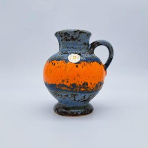 Op de afbeelding: Een vintage keramische kan met een handvat. De kan heeft een blauw en oranje kleurenschema met een donkere basis. Er is een kleine sticker op de voorkant bevestigd. De kan wordt waarschijnlijk gebruikt voor decoratieve doeleinden.