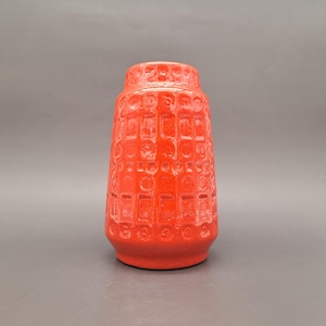 May include: A bright red ceramic vase with a textured, geometric design. The vase has a cylindrical form with a gently widening top and a solid base. The surface features a repeating grid of rectangular and circular indentations.