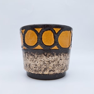 May include: A ceramic planter with a geometric design. The top section features a pattern of orange circles outlined in black. The middle section has a speckled, beige and brown texture, and the base is dark brown.