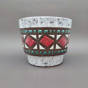 Peut inclure: Un pot de fleurs en céramique avec un extérieur blanc moucheté et une bande marron ornée de losanges rouges et de carrés turquoise. Le pot a un dessus large et légèrement évasé et une base plus petite.
