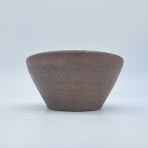 May include: A small, round ceramic bowl with a tapered base and a wide, open top. The exterior surface features a textured, matte finish in earthy shades of brown and dark grey, displaying a mottled, speckled appearance throughout the clay body.