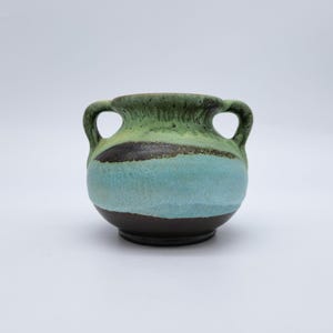 Pode incluir: Um pequeno vaso de cerâmica com duas alças. O vaso apresenta uma base castanha escura, uma faixa turquesa e um topo verde com uma faixa castanha escura. O vaso tem uma superfície texturizada e um aspeto rústico.