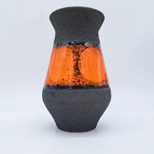 Puede incluir: Un jarrón de cerámica con base y borde negros texturizados, y una sección central naranja vibrante. La sección naranja tiene un patrón moteado, creando un contraste llamativo con el negro.