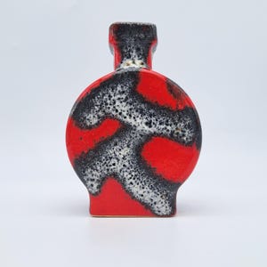 May include: A ceramic vase with a flat, circular body and a short neck. The surface features a vibrant red glaze contrasted by an abstract, textured pattern of black and white speckles that swirl across the front of the vessel.