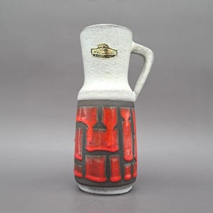 Puede incluir: Un jarrón de cerámica con la parte superior blanca y un diseño texturizado rojo y negro. El jarrón tiene un asa y una etiqueta que dice "Bay Keramik Handgemalt". El jarrón mide aproximadamente 20 cm de alto.