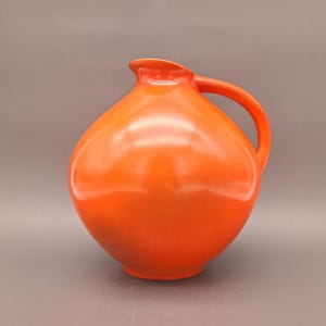 Puede incluir: Una jarra de cerámica naranja brillante con cuerpo redondeado y una sola asa. La jarra tiene un acabado liso y brillante.