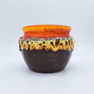 Op de afbeelding: Een keramische pot met een donkerbruine basis en een feloranje rand. De pot heeft een gestructureerd, gelaagd ontwerp met gele en witte accenten. De pot heeft twee kleine handvatten aan de zijkanten.