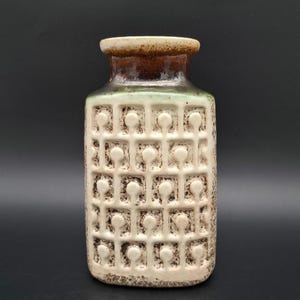 May include: A white ceramic vase with a brown and green glaze. The vase has a square shape with a raised geometric pattern of circles and squares.