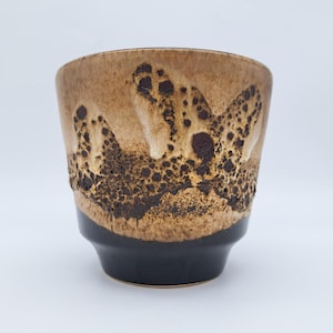Peut inclure: Une tasse en céramique avec un émail bicolore. La partie supérieure présente une finition texturée, tachetée de brun clair et foncé avec un aspect bullé et cratérisé, tandis que la partie inférieure est noir mat.